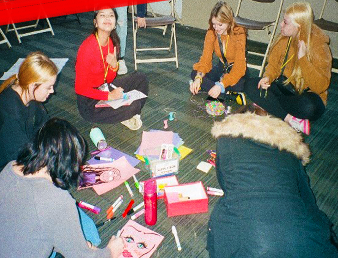 Group of young people sitting on the floor in a circle, working on art projects with markers, paper, and other supplies.